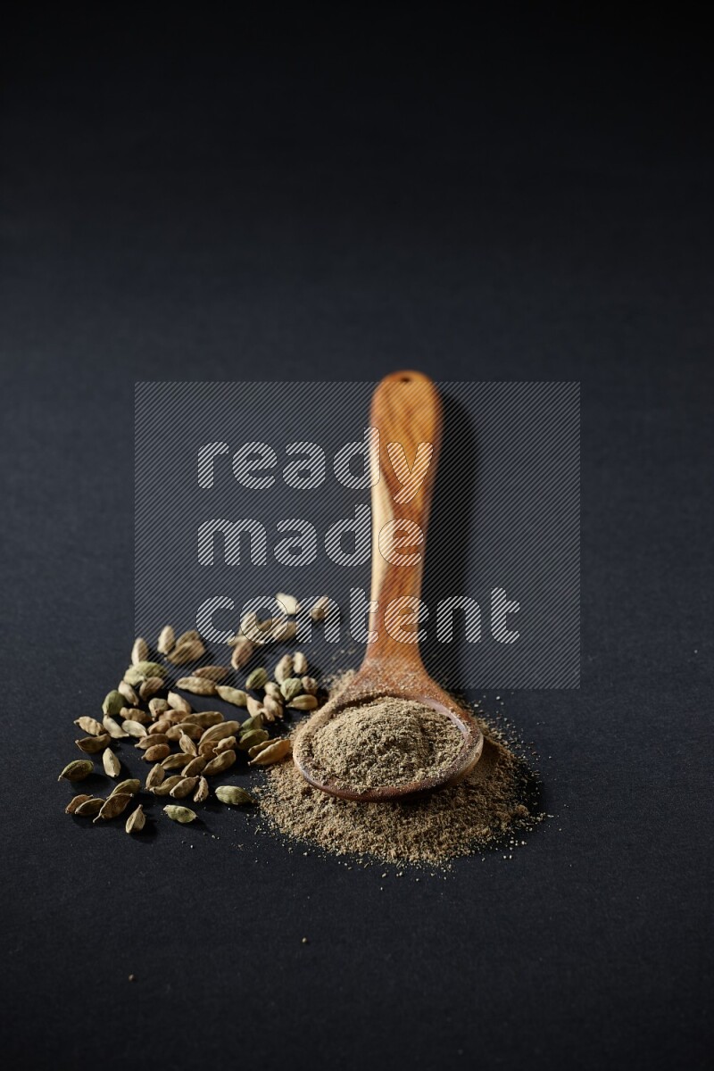 A wooden ladle full of cardamom powder and cardamom seeds beside it on black flooring