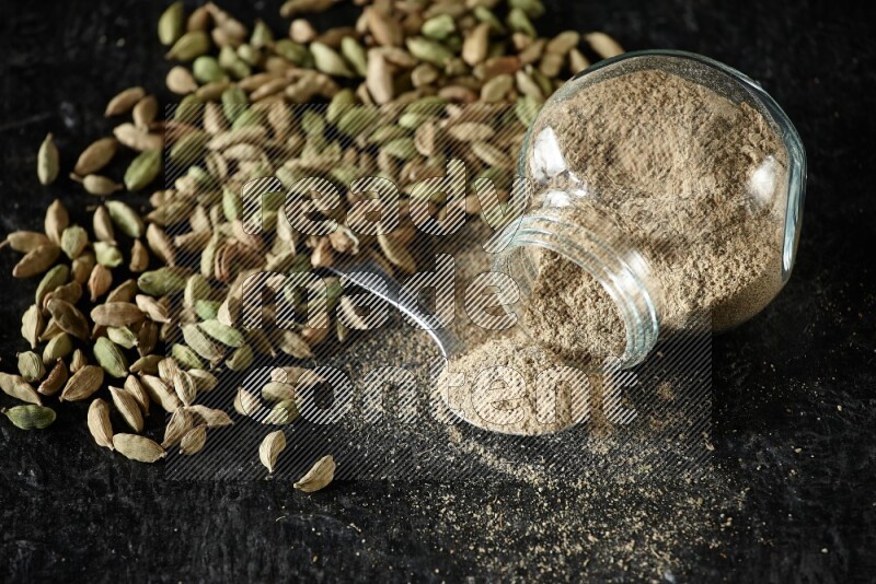 A flipped glass spice jar and a metal spoon full of cardamom powder and cardamom seeds spreaded on textured black flooring