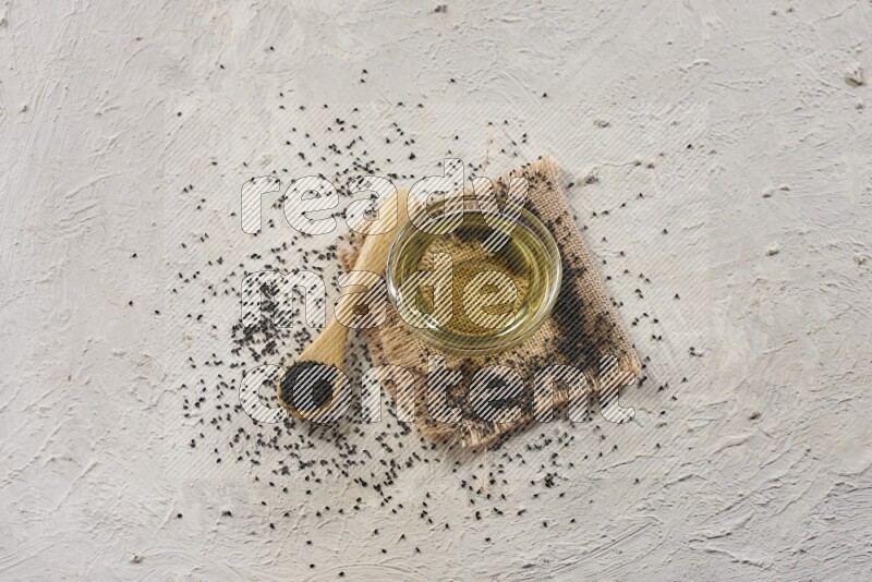 وعاء زجاجي ممتلئ بزيت حبة البركة وملعقة خشبية مليئة ببذور حبة البركة مع بعض البذور المتناثرة علي قطعة من القماش على أرضية بيضاء