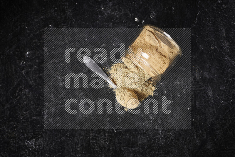 A glass jar full of ground ginger powder flipped with some spilling powder on black background