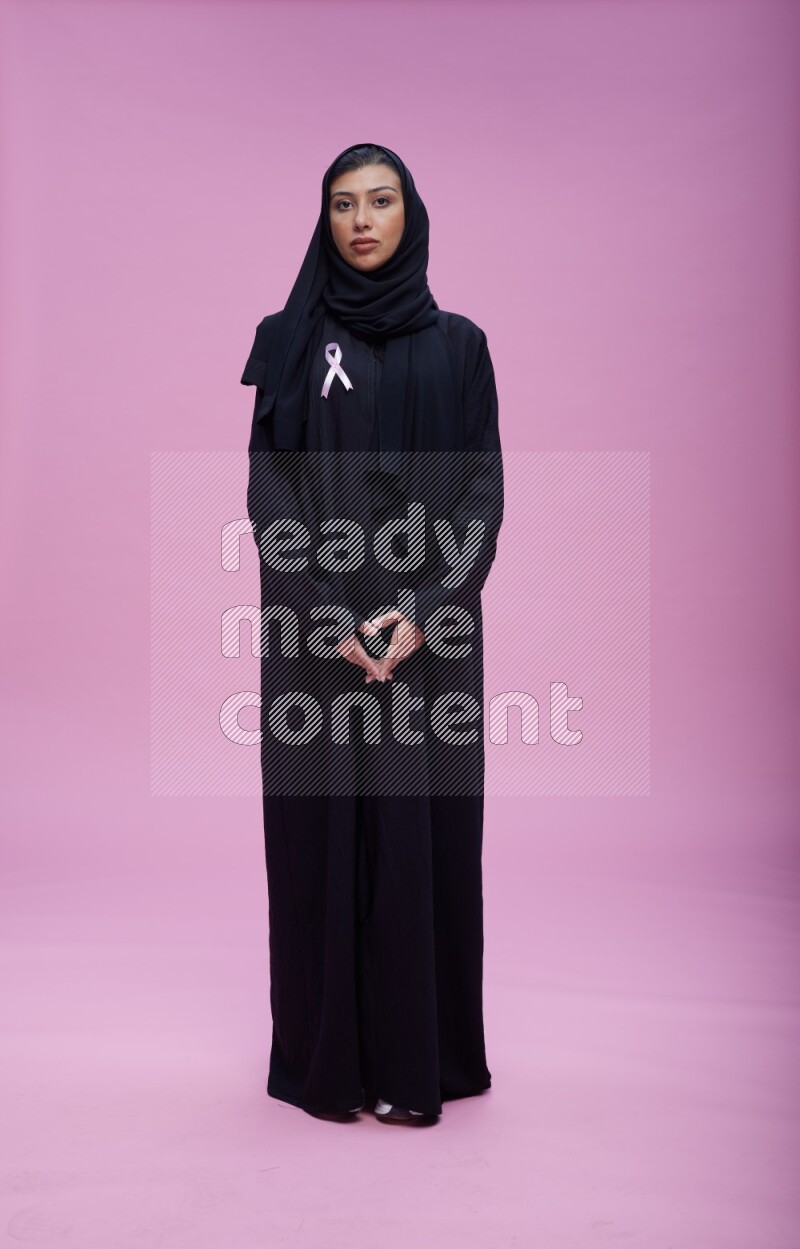 Saudi woman wearing pink ribbon on Abaya standing interacting with the camera on pink background