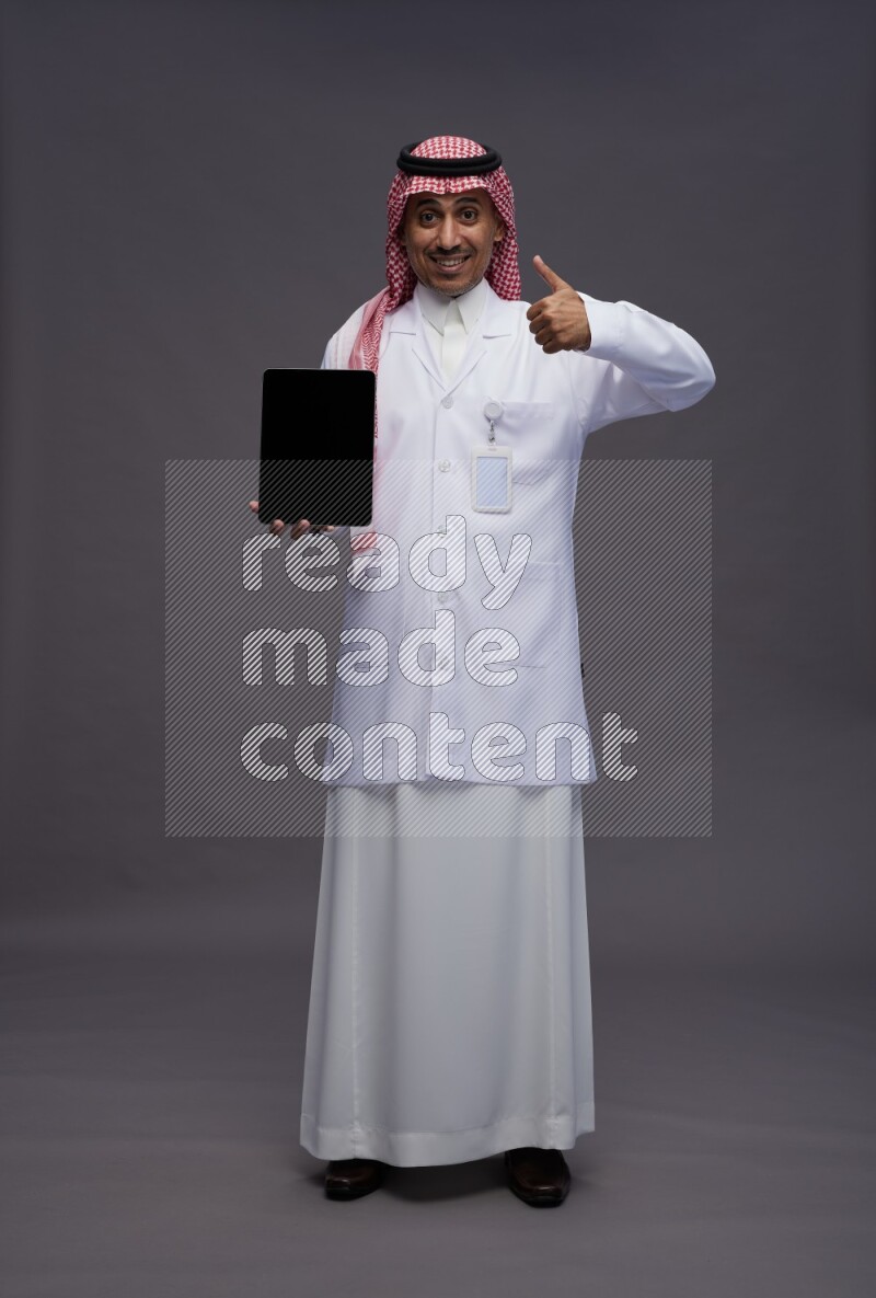 Saudi man wearing thob with lab coat and shomag with pocket employee badge standing showing tablet to camera on gray background