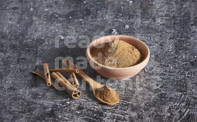 wooden bowl full of cinnamon powder and a wooden spoon full of it with cinnamon sticks on a textured black background