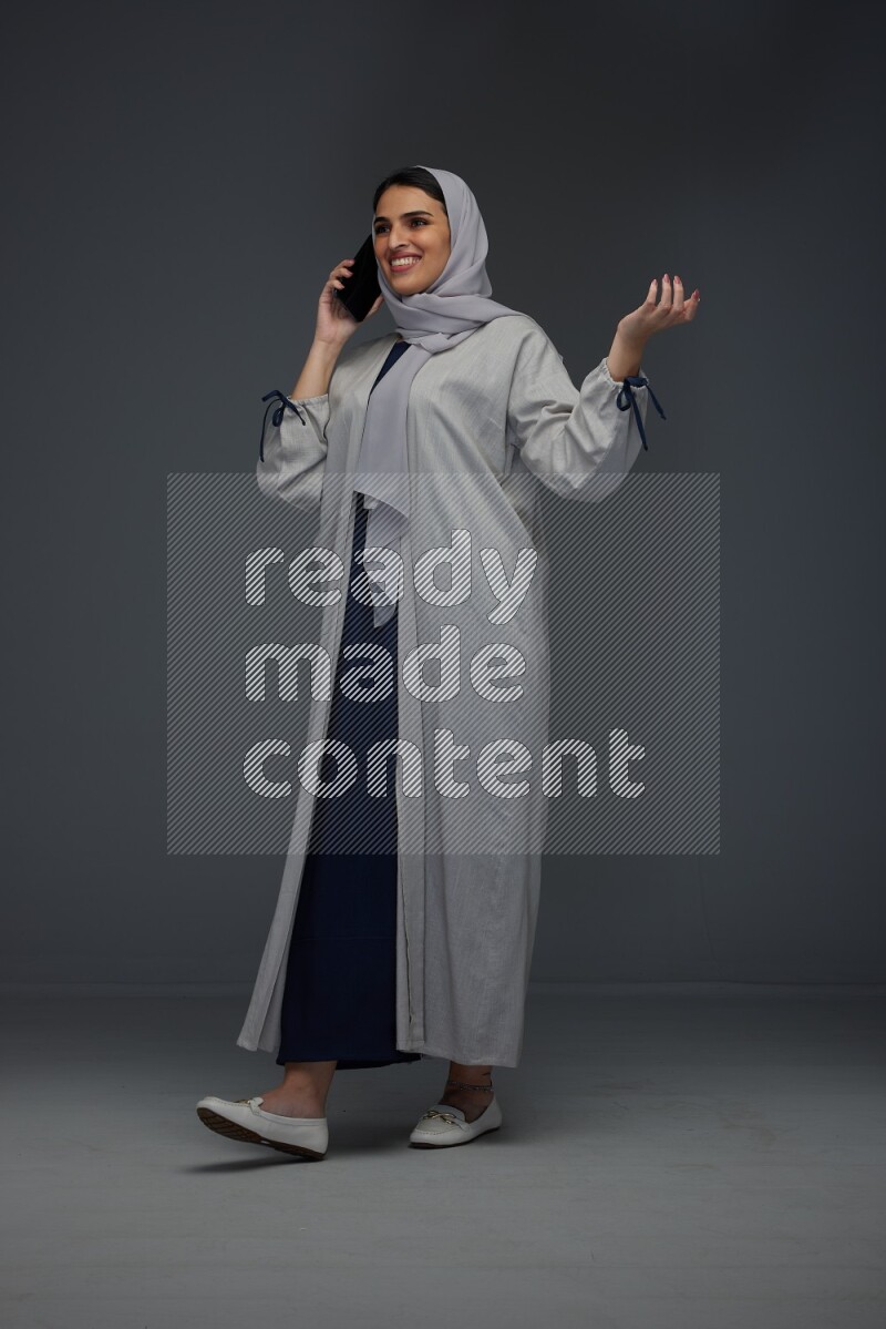 A Saudi woman wearing a light gray Abaya and head scarf standing and talking in the phone eye level on a grey background