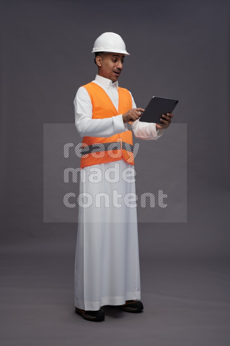 Saudi man wearing thob with engineer vest standing working on tablet on gray background