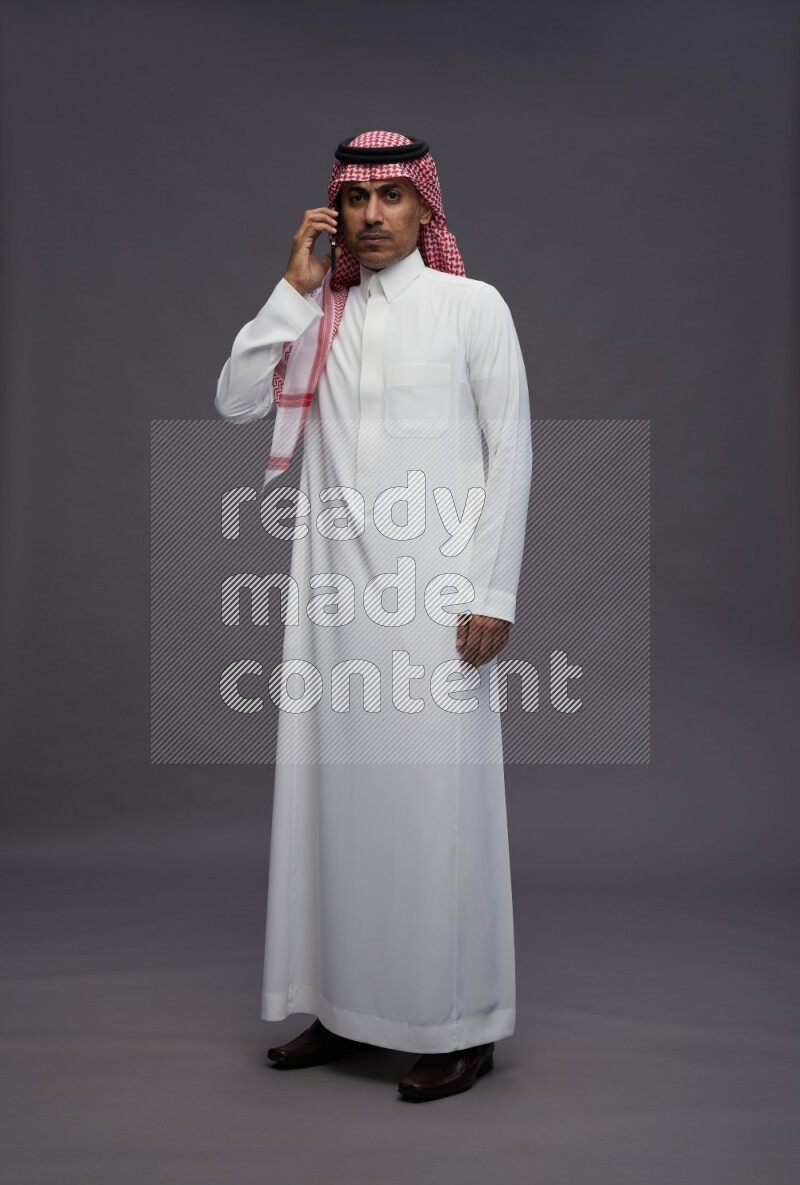 Saudi man wearing thob and shomag standing talking on phone on gray background