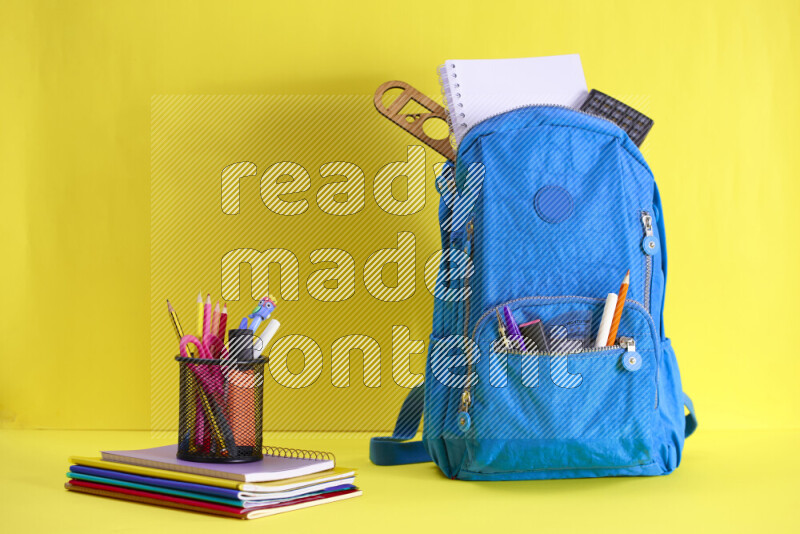 A school bag with assorted school supplies in and beside it on yellow background