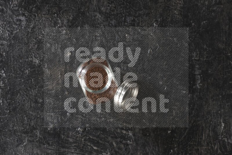 A glass spice jar full of cloves powder on textured black flooring