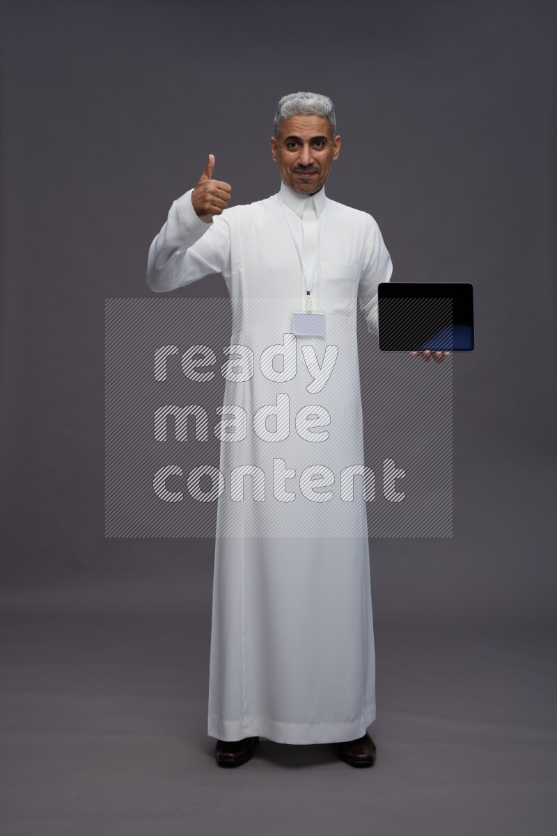 Saudi man wearing thob with neck strap employee badge standing showing tablet to camera on gray background