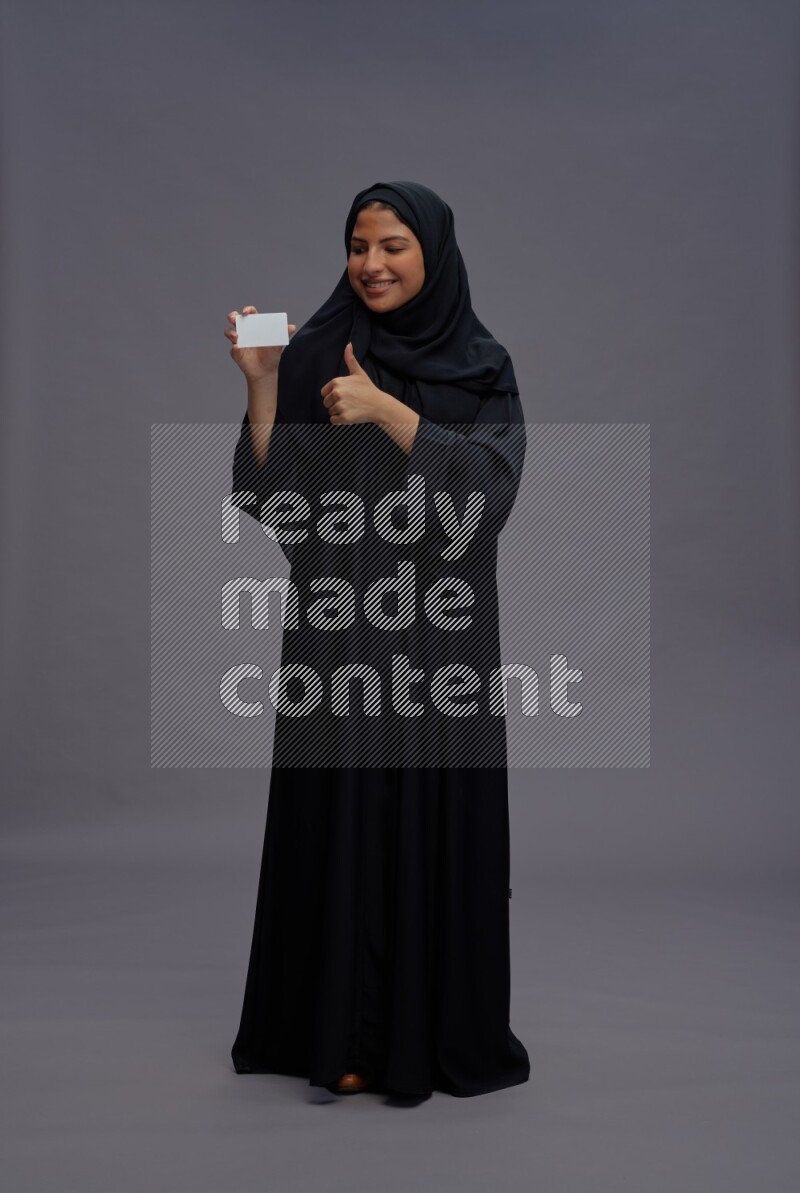 Saudi woman wearing Abaya standing holding ATM card on gray background