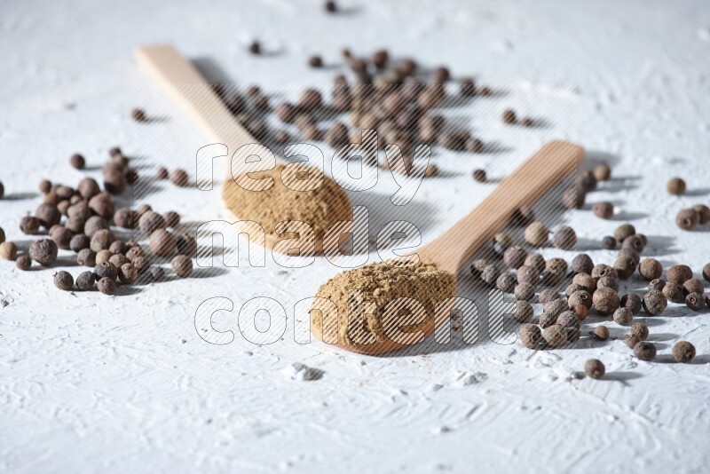 Wooden spoons full of allspice powder and allspice whole balls spreaded on a textured white flooring