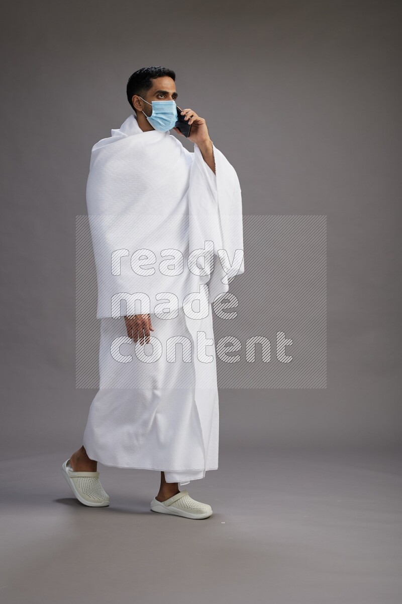 A man wearing Ehram with face mask Standing talking on phone on gray background