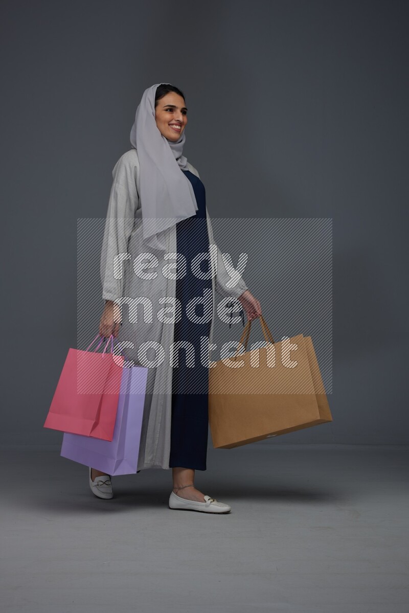A Saudi woman wearing a light gray Abaya and head scarf standing and holding shopping bags on a grey background