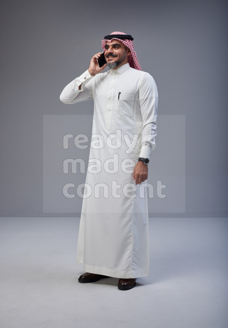 Saudi man Wearing Thob and red Shomag standing talking on phone on Gray background
