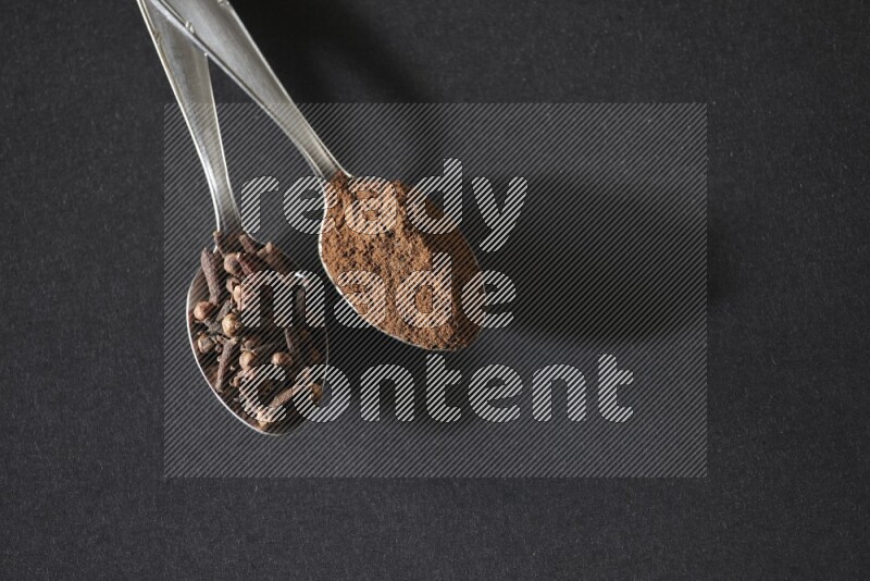 2 metal spoons, one full of cloves grains and the other full of cloves powder on a black flooring