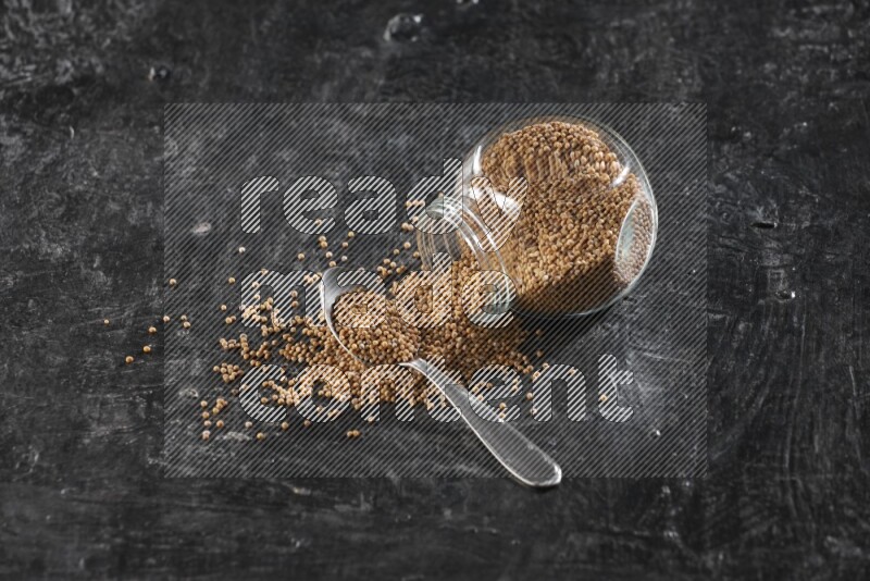 A glass spice jar and a metal spoon full of mustard seeds and jar is flipped with fallen seeds on a textured black flooring