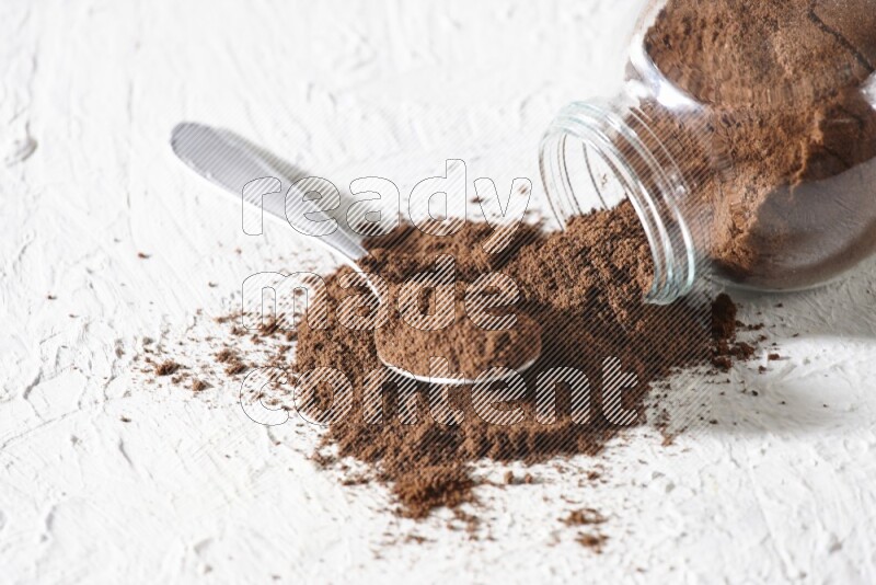 A flipped glass spice jar and a metal spoon full of cloves powder and powder came out of the jar on textured white flooring