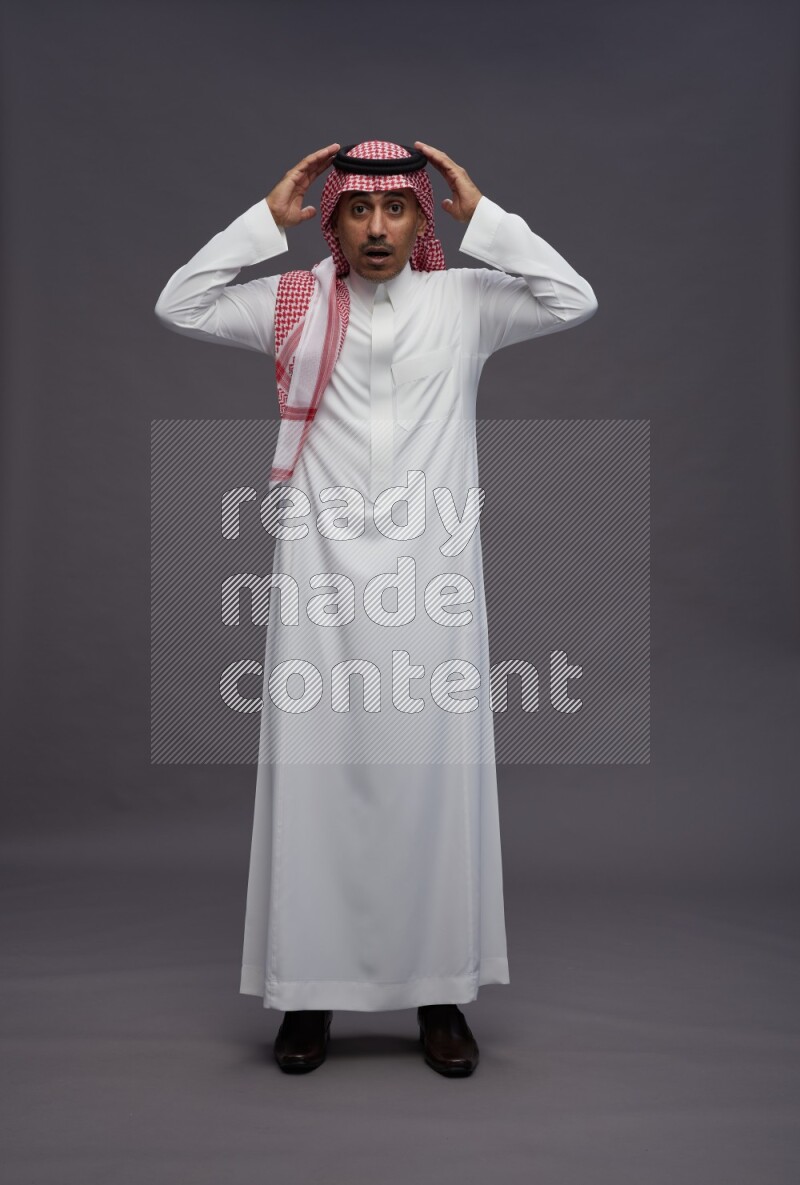 Saudi man wearing thob and shomag standing hands behind head on gray background