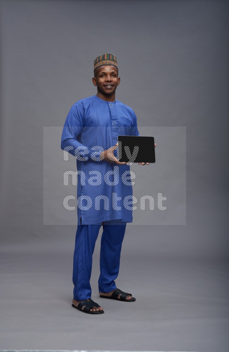 Man wearing Nigerian outfit standing showing tablet to camera on gray background