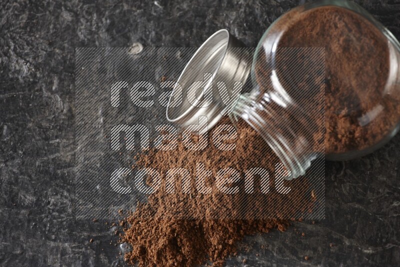 A flipped glass spice jar full of cloves powder and powder came out of it on textured black flooring