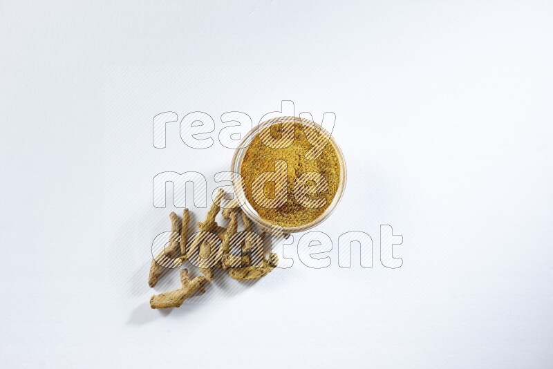 A glass bowl full of turmeric powder and dried whole fingers beneath it on white flooring