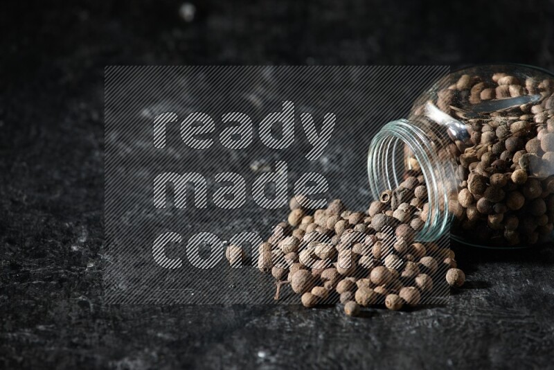 وعاء زجاجي للتوابل مقلوب ممتلئ بحبوب بهار الفلفل الحلو والحبوب تناثرت منه على خلفية سوداء