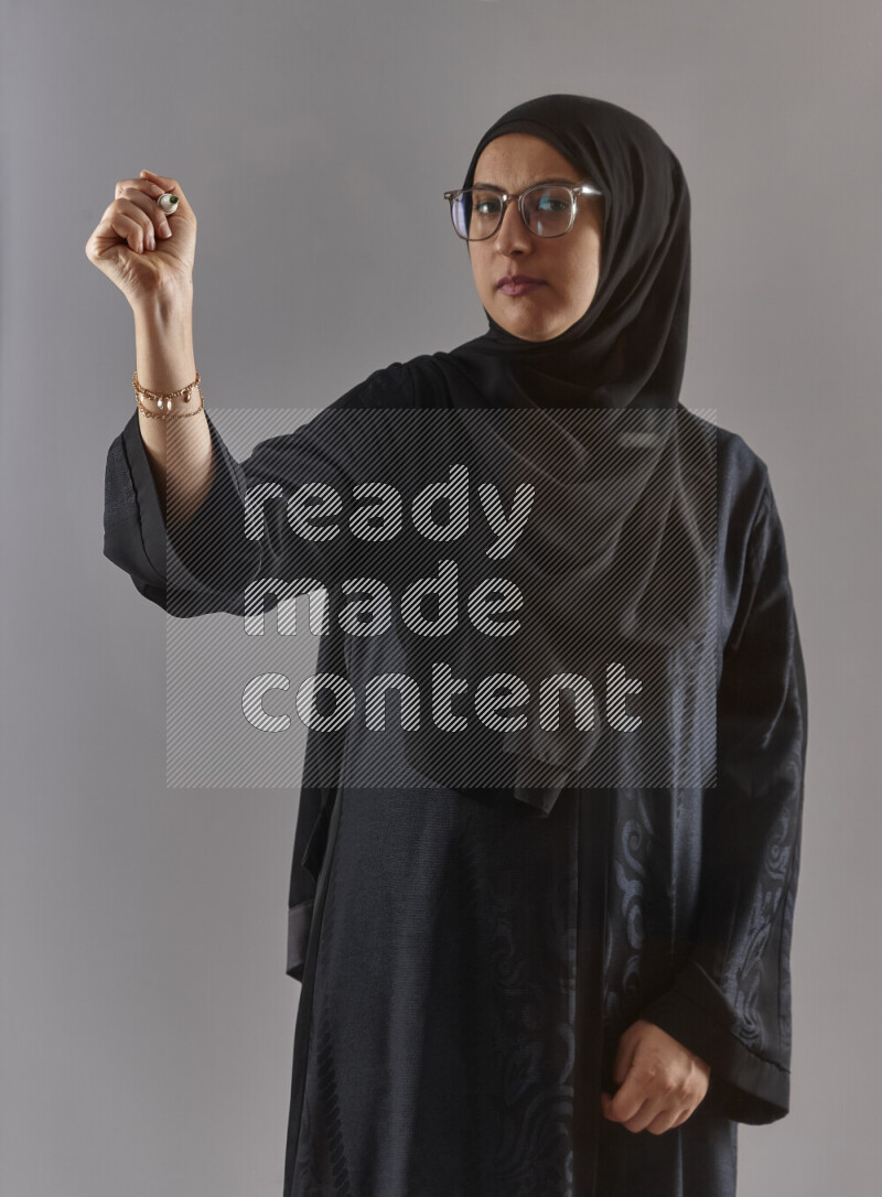 Woman in black abaya, hijab and glasses holding a marker pen to write on a transparent board with different reactions and expressions on grey background