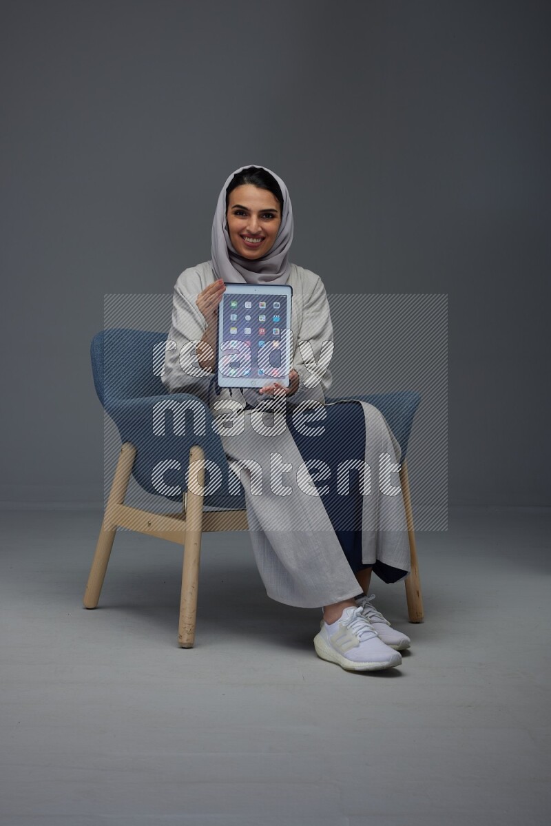 A Saudi woman wearing a light gray Abaya and head scarf sitting on a dark grey chair and showing the tablet's screen eye level on a grey background