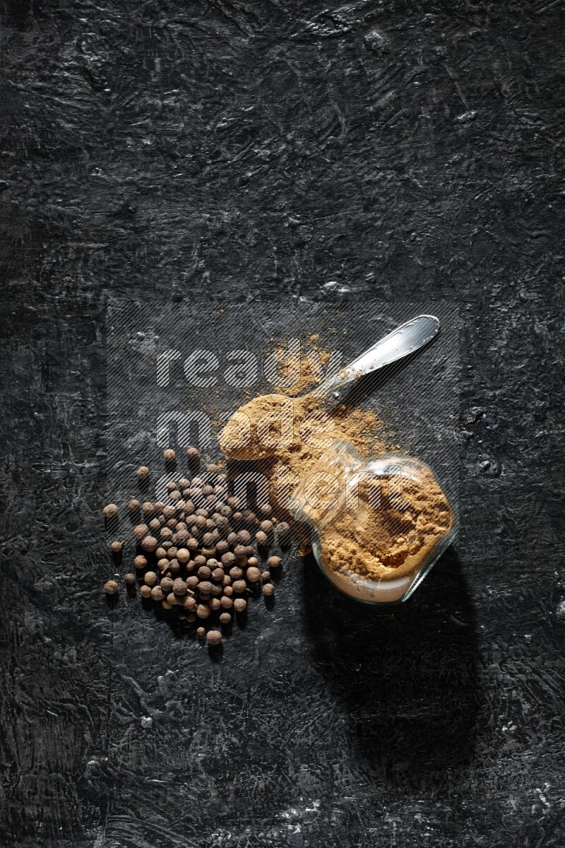 وعاء زجاجي للتوابل مقلوب وملعقة معدنية ممتلئان ببودرة بهار الفلفل الحلو والبودرة تناثرت من الوعاء مع بعض الحبوب على خلفية سوداء