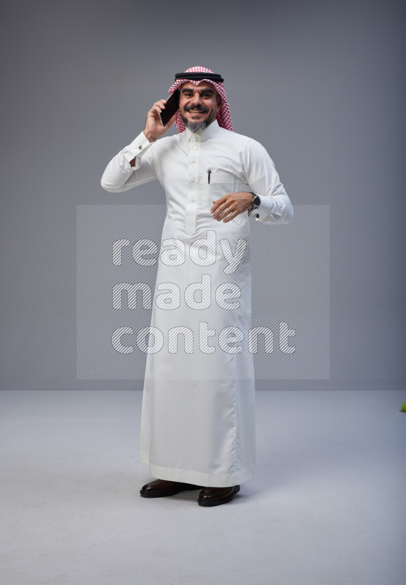 Saudi man Wearing Thob and red Shomag standing talking on phone on Gray background