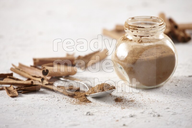 Herbal glass jar full cinnamon powder and a metal spoon surrounded by cinnamon sticks on a white background