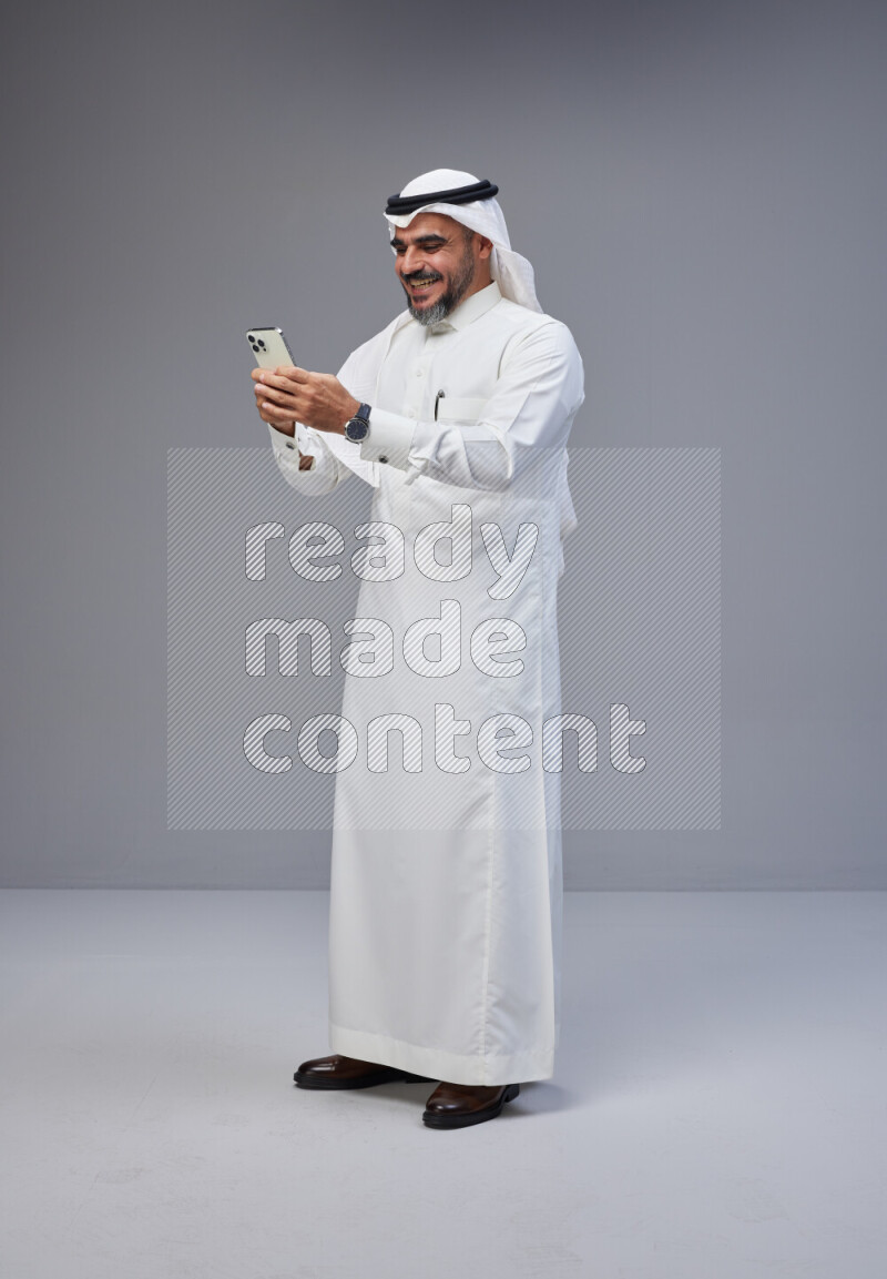 Saudi man Wearing Thob and white Shomag standing texting on phone on Gray background