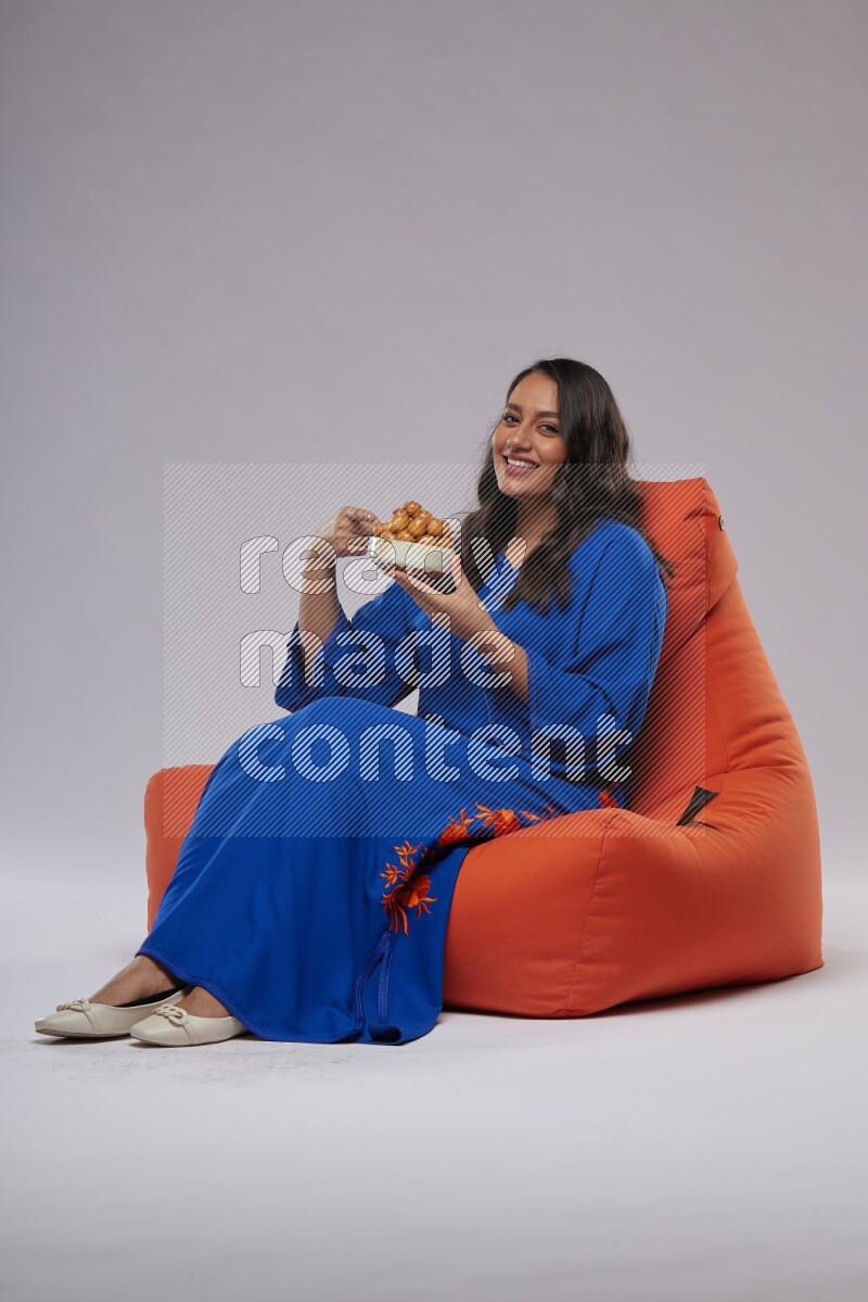 A Woman sitting on an orange beanbag wearing Jalabeya holding a plate of luqaimat