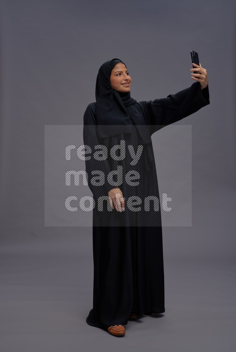 Saudi woman wearing Abaya standing taking selfie on gray background