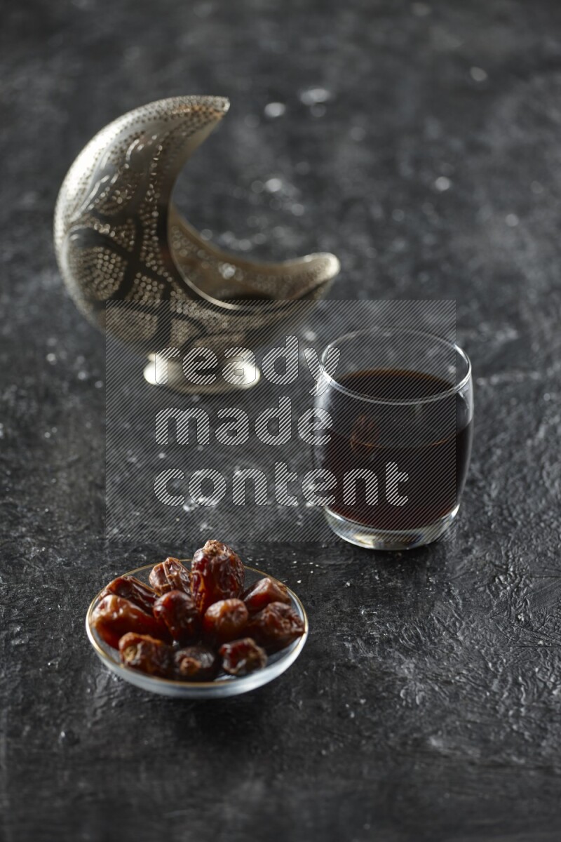 A silver lantern with different drinks, dates, nuts, prayer beads and quran on textured black background