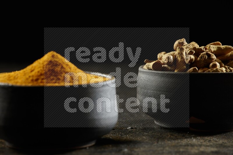 2 pottery black bowls, one full of turmeric powder and the other full of dried turmeric whole fingers on a textured black flooring