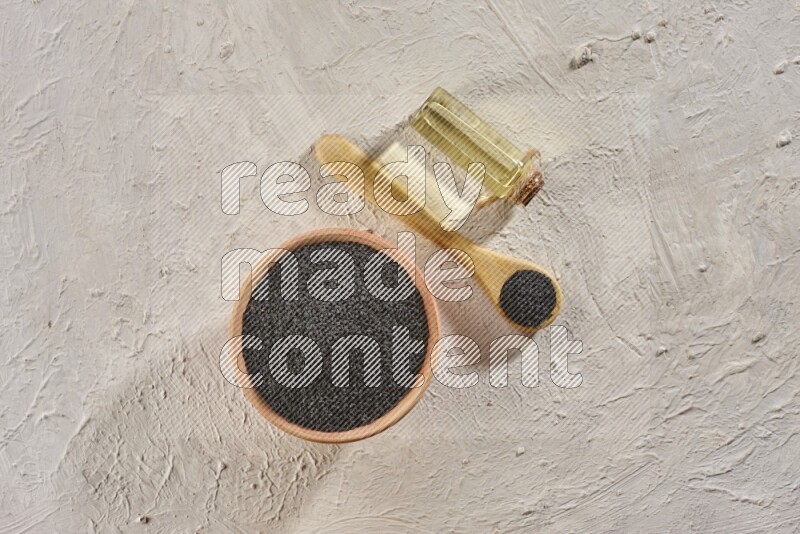 A wooden bowl and spoon full of black seeds with a bottle of black seeds oil on a textured white flooring