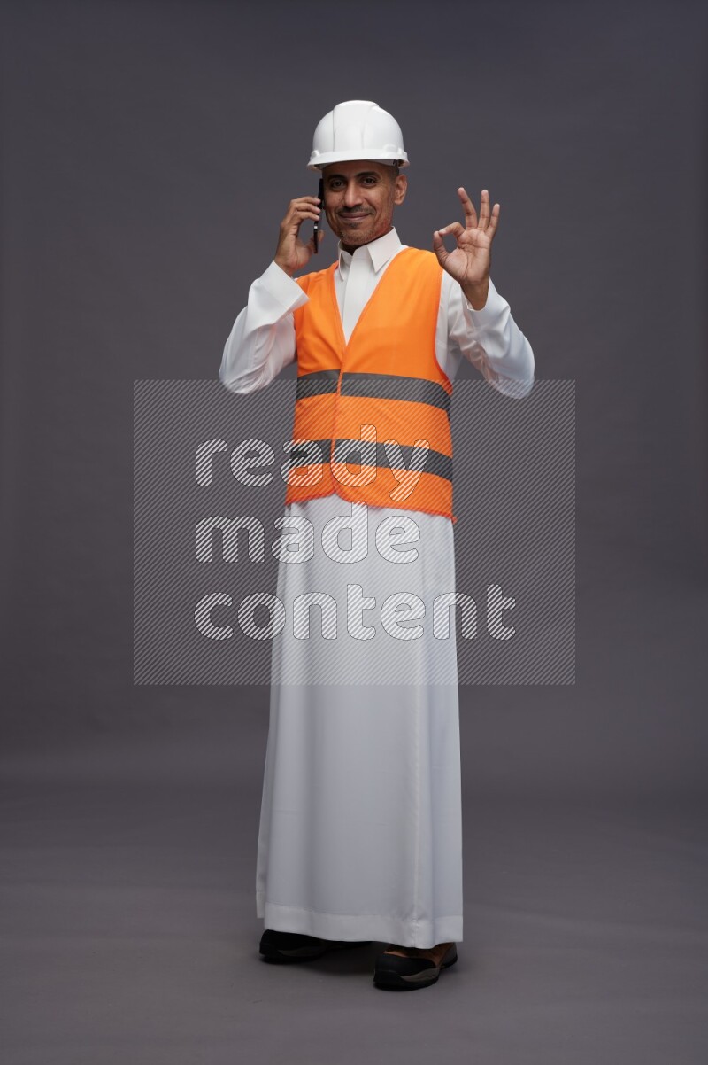Saudi man wearing thob with engineer vest standing talking on phone on gray background