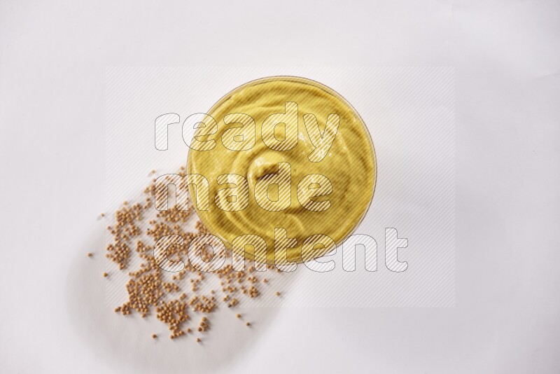 A glass bowl full of mustard paste with mustard seeds underneath on white flooring