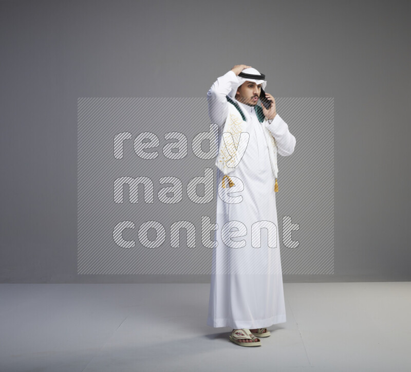 A saudi man standing wearing thob and white shomag with flag scarf on his neck talking on phone on gray background