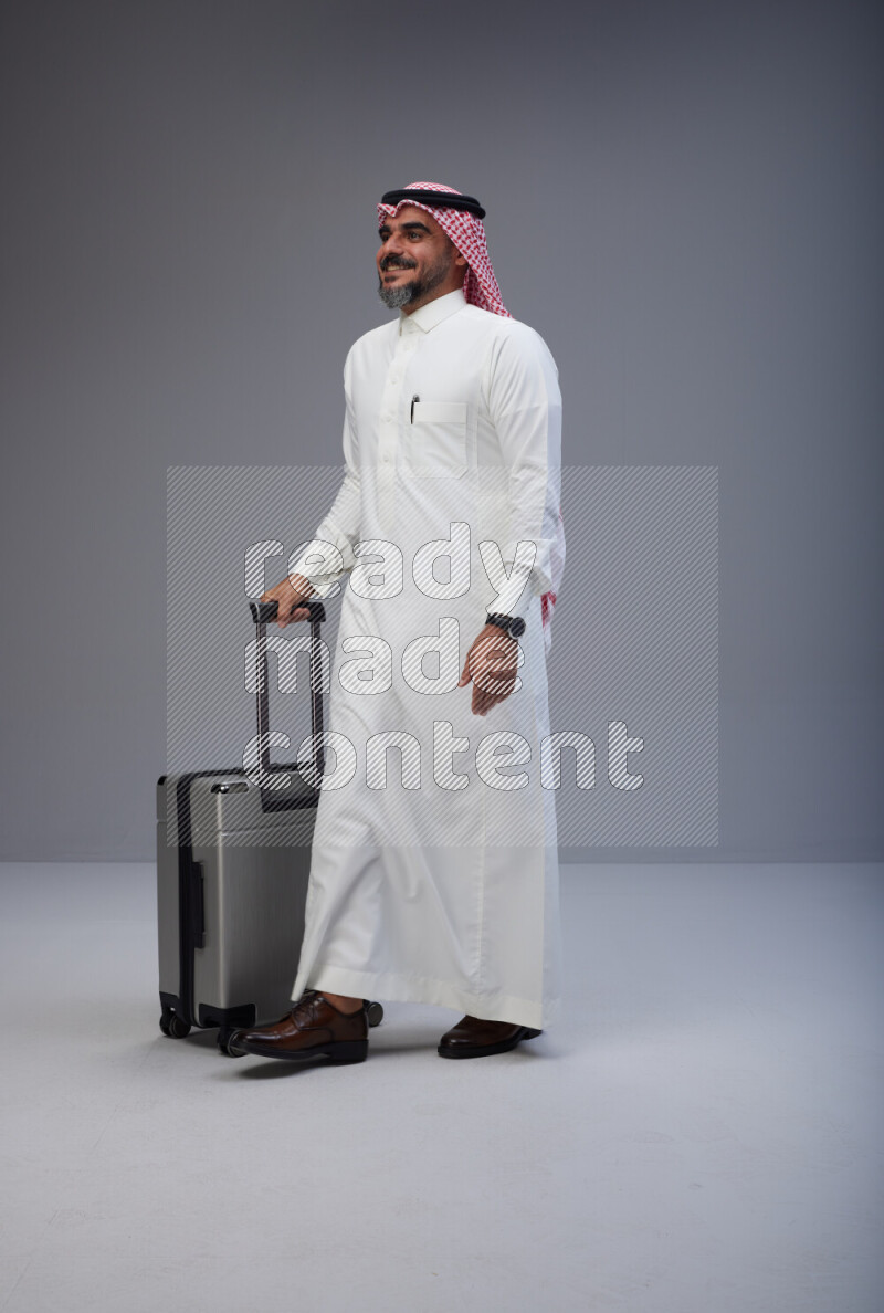 Saudi man wearing Thob and red Shomag standing holding Travel bag on Gray background
