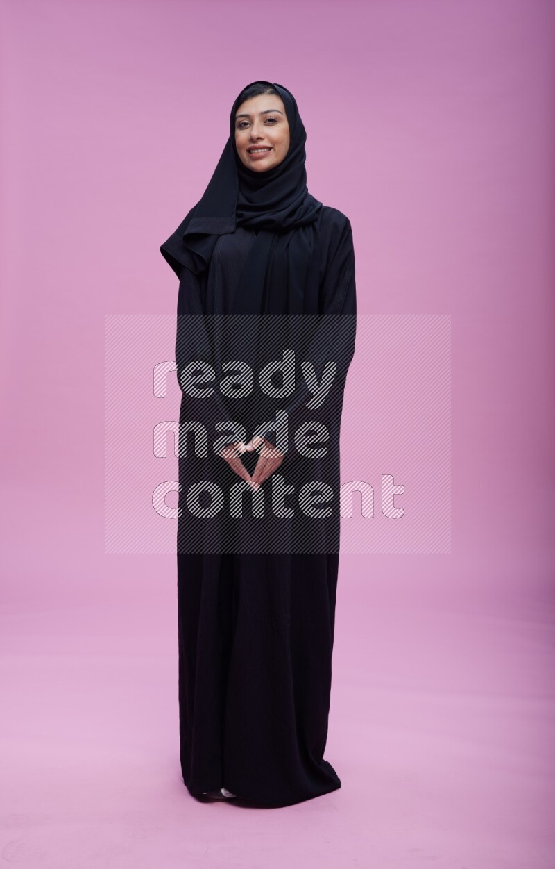 Saudi woman wearing Abaya standing interacting with the camera on pink background