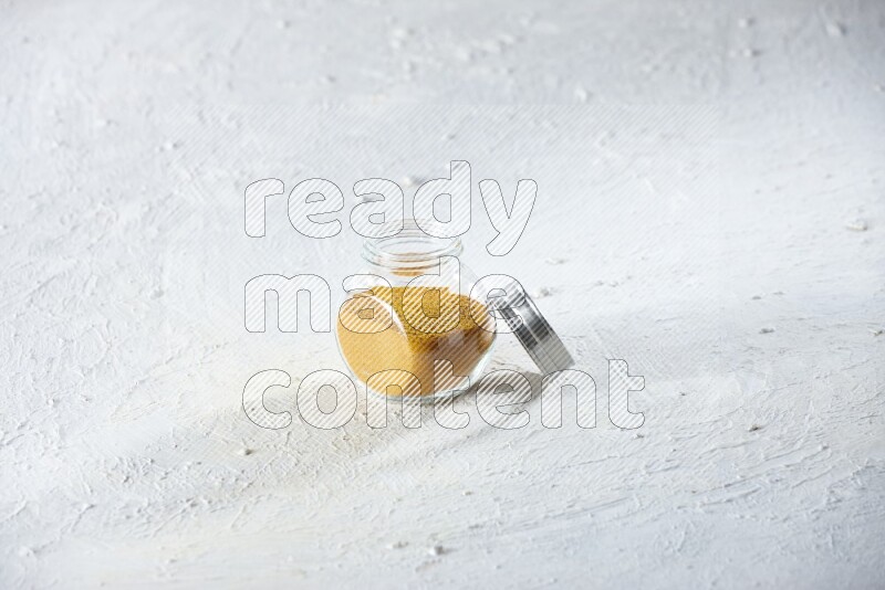 A glass spice jar full of turmeric powder on a textured white flooring