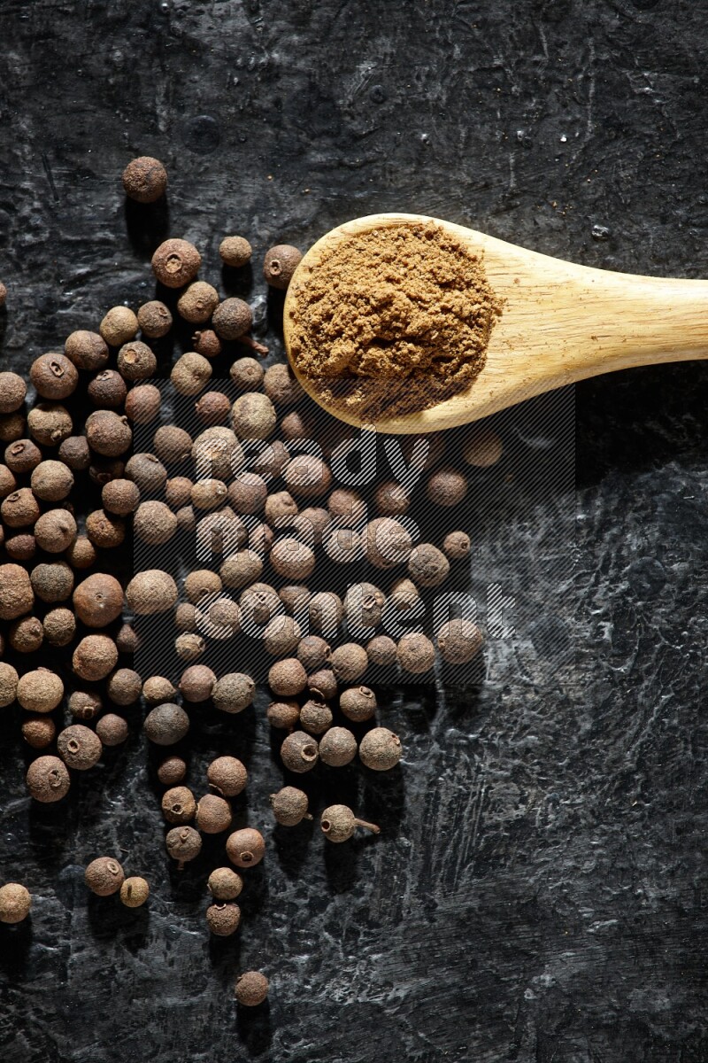A wooden spoon full of allspice powder and whole balls spreaded on a textured black flooring
