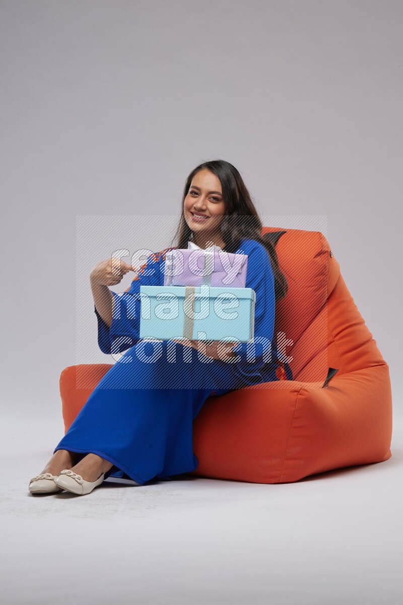 A woman sitting on an orange beanbag wearing Jalabeya holding a gift box