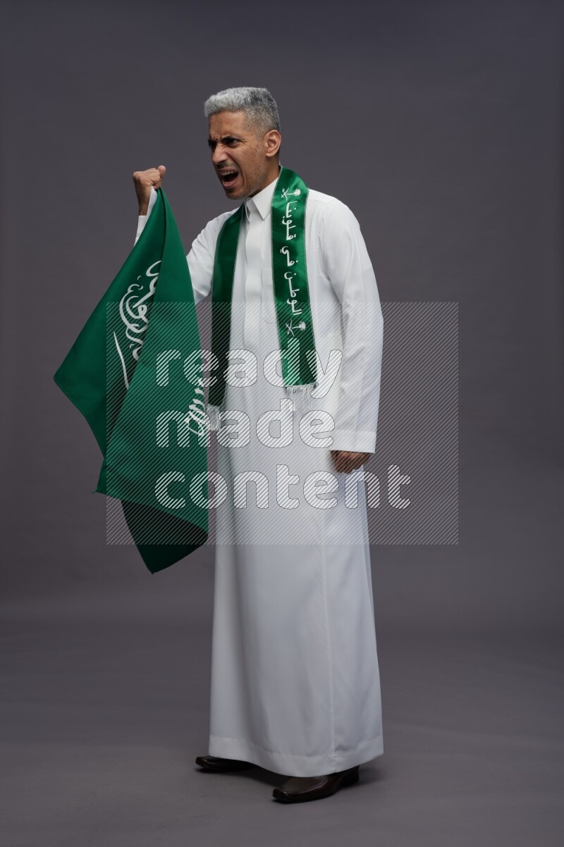 Saudi man wearing thob standing holding Saudi flag on gray background
