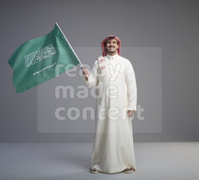 A Saudi man standing wearing thob and red shomag raising big Saudi flag on gray background