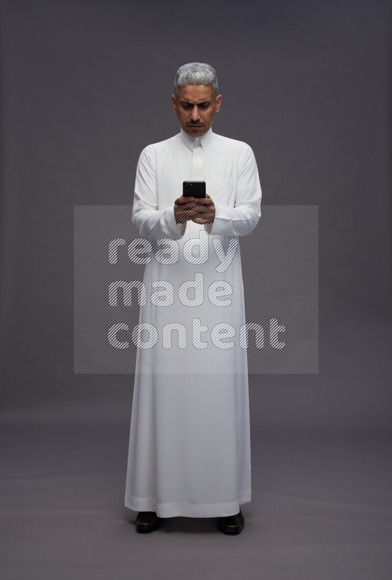 Saudi man wearing thob standing texting on phone on gray background
