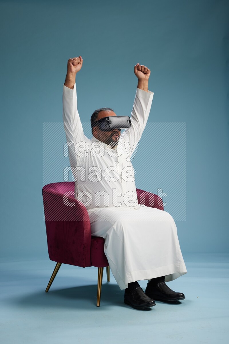 Saudi Man without shimag sitting on chair  gaming with VR on blue background