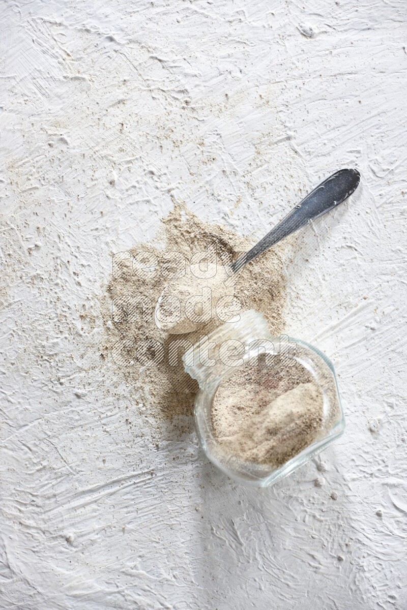 A flipped herbal glass jar and metal spoon full of white pepper powder with spilled powder on textured white flooring